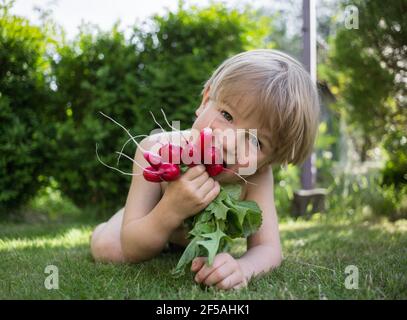 Happy cute Kleinkind Junge liegt auf grünem Gras mit einer Ernte von Radieschen aus dem Gartenbett. Kleiner Helfer, Landwirtschaft, gesunde Ernährung. Kindheit in den s Stockfoto