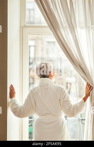 Rückansicht einer älteren Frau im Bademantel, der Vorhänge öffnet Stockfoto