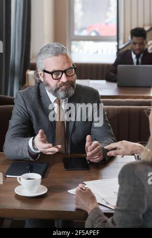 Bärtiger reifer Geschäftsmann, der mit seinem Kollegen am Tisch sitzt Und die Arbeit bei der Besprechung im Restaurant besprechen Stockfoto