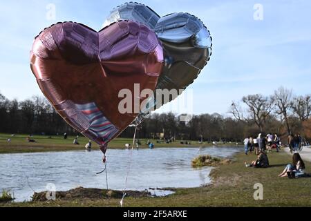 25. März 2021, Bayern, München: Im Englischen Garten hängen zwei herzförmige Luftballons an Schnüren, damit sie nicht wegfliegen, während Jugendliche und Kinderwagen das warme Frühlingswetter im Park genießen. Foto: Katrin Requadt/dpa Stockfoto
