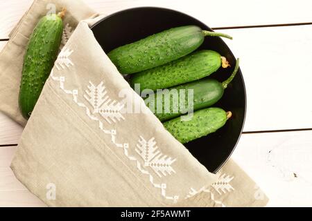 Mehrere reife dunkelgrüne Bio-Gurken mit einer Leinenserviette und einem schwarzen Keramikteller, auf einer weißen Holzplatte. Stockfoto