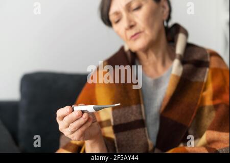 Ältere Frau, die die Körpertemperatur überprüft, auf der mit einer Decke bedeckten Couch liegt, sich krank fühlt, an Fieber leidet Stockfoto