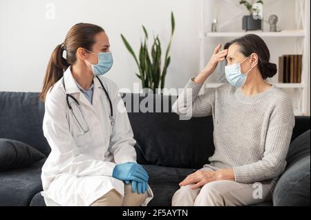 Krankenschwester in Maske Pflege von älteren weiblichen Patienten trägt Maske zu Hause während Coronavirus Pandemie. Eine reife Frau beklagt sich beim Arzt wegen Unwohlsein, Unwohlsein. Arzttermin zu Hause Stockfoto