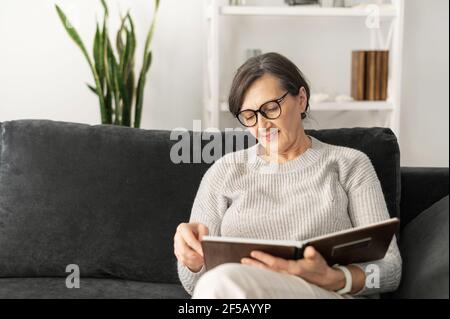 Pensionierte Frau verbringt Freizeit mit dem Betrachten Fotoalbum, ältere Frau sitzt auf der gemütlichen Couch zu Hause, erinnern Jugend, beobachten alte Fotos. Großmutter schaut durch Bilder von Enkeln Stockfoto