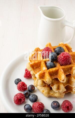 Draufsicht auf gestapelte Waffeln mit Himbeeren und Heidelbeeren, auf weißem Tisch mit weißem Krug, im Portrait Stockfoto