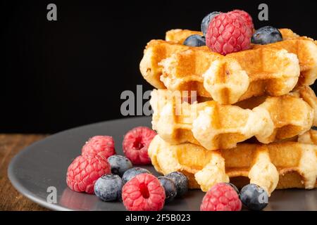 Nahaufnahme von gestapelten Waffeln mit Himbeeren und Heidelbeeren auf dunklem Teller, auf Holztisch, schwarzer Hintergrund, horizontal Stockfoto