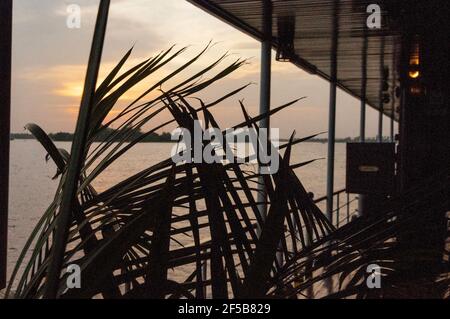 Abendstimmung auf dem Mekong. Blick durch ein Palmblatt auf die untergehende Sonne. Über die Uferfläche. Teilansicht eines Flussbootes. Stockfoto