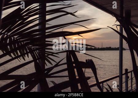 Abendstimmung auf dem Mekong. Blick durch ein Palmblatt auf die untergehende Sonne. Über die Uferfläche. Stockfoto