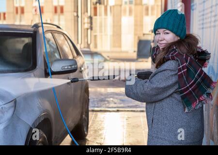 Lächelnde kaukasische Frau in warmer Kleidung waschen ihr suv-Auto an selbst Auto-Waschstation im Freien, mit Hochdruck-Wasserstrahl Stockfoto