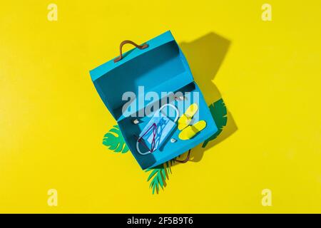 Reisen während Corona Virus Epidemie Konzept. Schutzmaske, Sonnenbrille und Flip Flops in blauem Koffer auf gelb Stockfoto