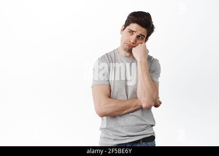 Trauriger und düsterer erwachsener Mann, der sich verärgert fühlt, den Kopf an der Hand lehnt und mit einem düsteren Gesicht zur Seite schaut, vor weißem Hintergrund verzweifelt steht Stockfoto