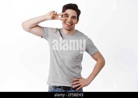 Glücklicher und positiver junger Mann, der Friedenszeichen zeigt, mit V-Zeichen lächelt und fröhlich aussieht, auf weißem Hintergrund steht Stockfoto