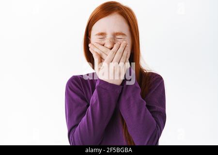 Portrait von niedlichen Rotschopf Mädchen niesen, bedeckt ihren Mund und Falten Nase, stehen über weißem Hintergrund. Kind versteckt ihr Lächeln, lachend und scherzend Stockfoto