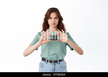 Traurige junge Frau in T-Shirt zeigt ihre Kunststoff-Kreditkarte und grimacing aufgeregt, schlechte Laune, stehen enttäuscht vor weißem Hintergrund Stockfoto