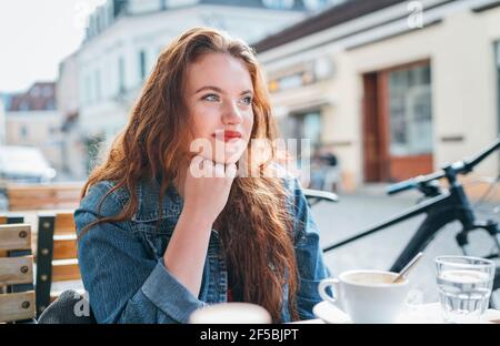 Porträt der traurigen roten gewellten langen Haar kaukasischen Frau sitzt auf einer gemütlichen Terrasse im Freien Café auf der Straße und Blick auf die Straße. Junge Frau takin Stockfoto