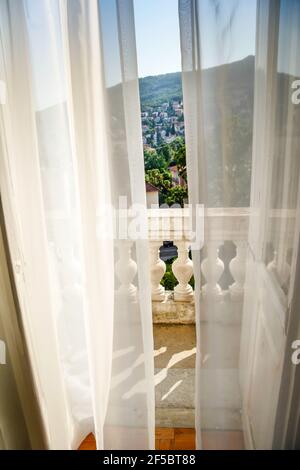 Ein leichter Wind bläst bodenlange Gardinen in einem Romantische Szene von einem offenen Satz von französischen Türen auf Ein Balkon mit einem Blick auf die Vorberge und Häuser Stockfoto