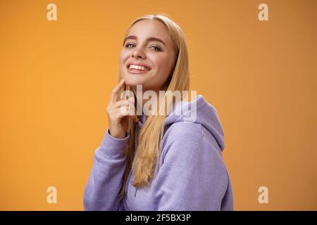 Attraktive freche selbstbewusste kaukasische blonde Frau in lila Kapuzenpullover Drehen Kamera berühren Kinn frech Lächeln Kamera überprüfen Sie interessante Sache Stockfoto