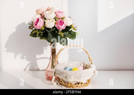 Ostern bunte Eier in busket auf weißem Holztisch. Bouquet von Rosenblüten auf dem Hintergrund. Frohe Ostern. Frühlingsstimmung. Stockfoto