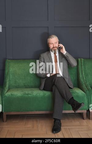 Reifer eleganter Geschäftsmann sitzt auf dem modernen Sofa und spricht weiter Handy im Zimmer Stockfoto