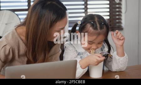 Asiatische Familie junge Mutter und ihre kleine Tochter lächeln mit Laptop-Computer zusammen für online lernen und Instant Nudeln zu Hause essen, Kind EA Stockfoto