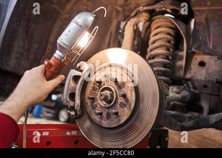 Ein Automechaniker inspiziert Bremsscheiben und Bremsbeläge mit einer Taschenlampe. Auto auf einem Reparaturständer. Technische Servicestation für Auto. Stockfoto