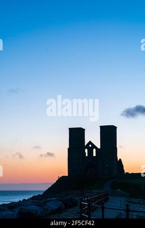 Reculver Silhouette des 12. Jahrhunderts Zwillingsstürme am Meer an der Küste von Kent, silhouettiert gegen den Morgenhimmel. Der Turm, eine bekannte Navigationshilfe für vorbeifahrende Schiffe, wurde der ursprünglichen angelsächsischen Kirche hinzugefügt, die auf dem Gelände gebaut wurde. Hauptsächlich klarer blauer Himmel mit gelbem und orangefarbenem Band am Horizont. Stockfoto