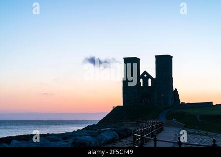 Reculver Silhouette des 12. Jahrhunderts Zwillingsstürme am Meer an der Küste von Kent, silhouettiert gegen den Morgenhimmel. Der Turm, eine bekannte Navigationshilfe für vorbeifahrende Schiffe, wurde der ursprünglichen angelsächsischen Kirche hinzugefügt, die auf dem Gelände gebaut wurde. Hauptsächlich klarer blauer Himmel mit gelbem und orangefarbenem Band am Horizont. Stockfoto