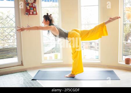 Junge Frau, die auf einem Bein in einem Krieger III steht Pose während ihres Yoga-Unterrichts zu Hause Stockfoto