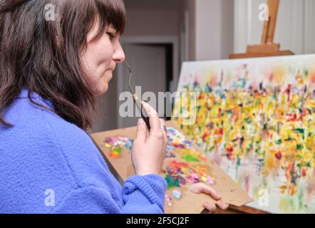 Junge Frau denkt und malt auf Leinwand mit Ölfarben zu Hause. Künstler Zeichnung auf Staffelei. Stockfoto