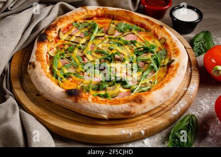 Hausgemachte Pizza mit Rucola, Gurken, Schinken und Soße auf einem hölzernen Pizzateller. Italienische Küche. Stockfoto