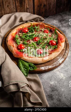 Italienische Küche. Dünne Pizza mit dicken Seiten. Pizza in Tomatensauce mit, Rucola, Käse, Kirschtomaten und Prosciutto. Hintergrundbild. Platz für Stockfoto