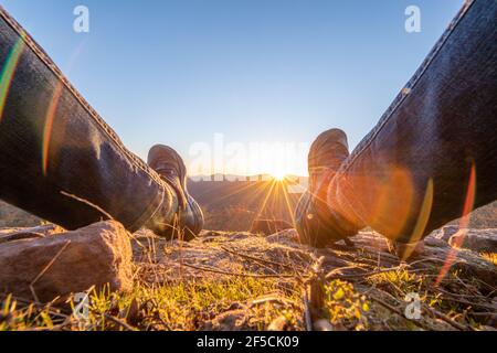 Die Sonne geht zwischen den Beinen eines Mannes unter Sitzen auf einem Berg Stockfoto