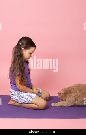 Eine rothaarige britische Katze streckt mit ihrer Pfote ein fünfjähriges Mädchen auf ihrem Schoß auf einer lila Sportmatte, isoliert auf einem rosa Hintergrund Stockfoto