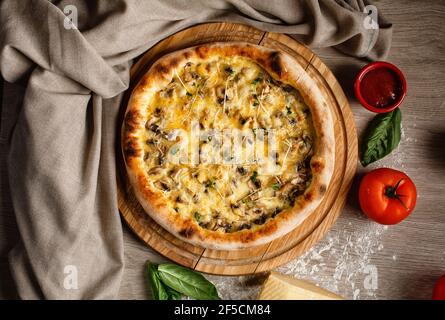 Heiße Pizza mit Pilzen und Käse auf einem runden Holzbrett. Pizza auf dem Tisch mit einem Stück Tuch, Mehl, Tomate, Sauce und Basilikumblättern dekoriert Stockfoto