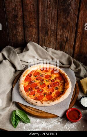 Frische italienische klassische original Pepperoni Pizza auf einem Holzbrett für Pizza. Gericht auf einem Tisch mit Basilikum, Tuch, Sauce und Mehl dekoriert. Foto für mich Stockfoto