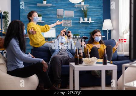Fröhliche fröhliche Frau schreien nach einem Gaming-Wettbewerb mit multiethnischen Freunden in zu Hause Wohnzimmer mit Gesichtsmaske verhindert Coronavirus Ausbreitung in Zeiten der globalen Pandemie. Stockfoto