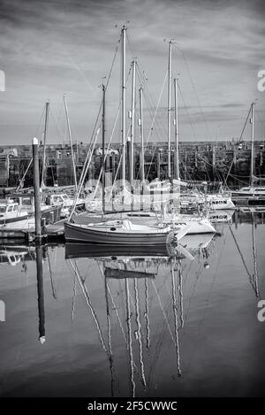Ein Jachthafen von Yachten liegt mit ihren Spiegelungen im ruhigen Wasser vor Anker. Ein Pier ist jenseits und ein Himmel mit Wolken oben. Stockfoto