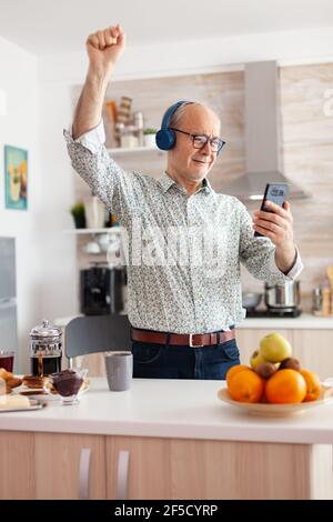 Fröhliche Erwachsene hören Musik tragen Kopfhörer in der Küche während des Frühstücks. Ältere Rentner genießen modernen Spaß glücklich Lebensstil, tanzen entspannt, lächelnd und mit moderner Technologie Stockfoto