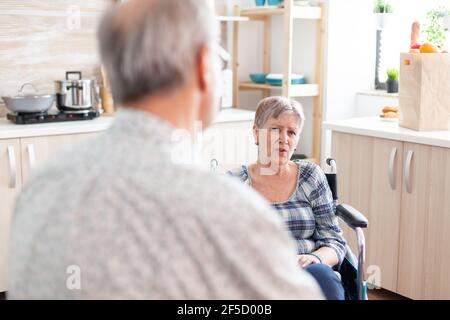 Besorgt behinderte ältere Frau im Rollstuhl im Gespräch mit Ehemann in der Küche sitzen. Altes Paar mit einem Gespräch über die Behandlung, Mann, der mit behinderten Menschen mit Gehbehinderungen lebt Stockfoto