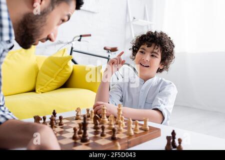 Fröhlicher Mulsim Junge zeigt mit Finger, während Schach spielen mit Vater auf verschwommenem Vordergrund Stockfoto