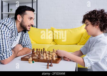 Lächelnder muslimischer Mann, der den Sohn beim Schachspielen zu Hause ansieht Stockfoto