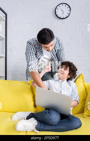 Lächelndes arabisches Kind, das auf dem Sofa mit Laptop sitzt und zeigt Mit Finger in der Nähe Vater Stockfoto