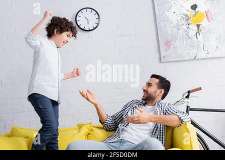 Fröhlicher arabischer Mann zeigt auf aufgeregt Sohn springen auf Sofa Mit geballten Fäusten Stockfoto