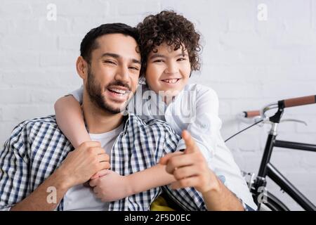 Fröhlicher arabischer Junge umarmt lachenden Vater zeigt mit dem Finger auf Kamera Stockfoto