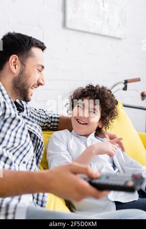 Fröhlicher arabischer Junge, der mit dem Finger in der Nähe des Vaters im Fernsehen zeigt Fernbedienung auf verschwommenem Vordergrund Stockfoto