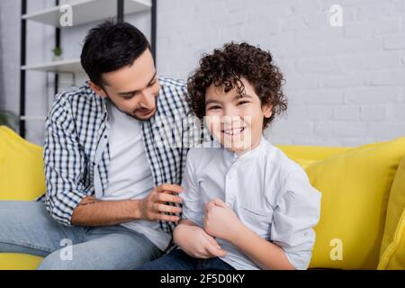 Fröhlicher arabischer Mann kitzelt fröhlichen Sohn, während er auf dem Sofa sitzt Zu Hause Stockfoto
