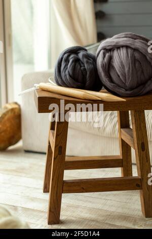 Zwei große Kugeln aus Merinowolle liegen auf einem Holz Hocker mit Holzstricknadeln vor dem Hintergrund eines Fenster und ein weißes Sofa Stockfoto