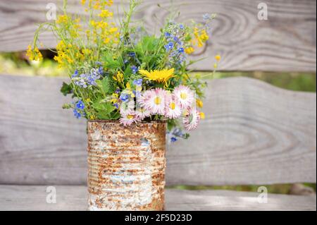 Ein Bouquet von Wildblumen aus Vergissmeinnicht, Gänseblümchen und gelben Löwenzahn in voller Blüte in einem rostigen rustikalen Glas vor einem Hintergrund von Holzbohlen in Stockfoto