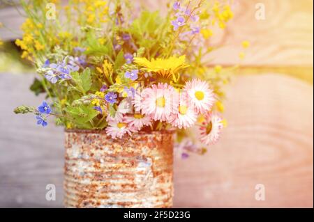 Ein Bouquet von Wildblumen aus Vergissmeinnicht, Gänseblümchen und gelben Löwenzahn in voller Blüte in einem rostigen rustikalen Glas vor einem Hintergrund von Holzbohlen in Stockfoto