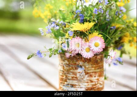 Ein Bouquet von Wildblumen aus Vergissmeinnicht, Gänseblümchen und gelben Löwenzahn in voller Blüte in einem rostigen rustikalen Glas vor einem Hintergrund von Holzbohlen in Stockfoto